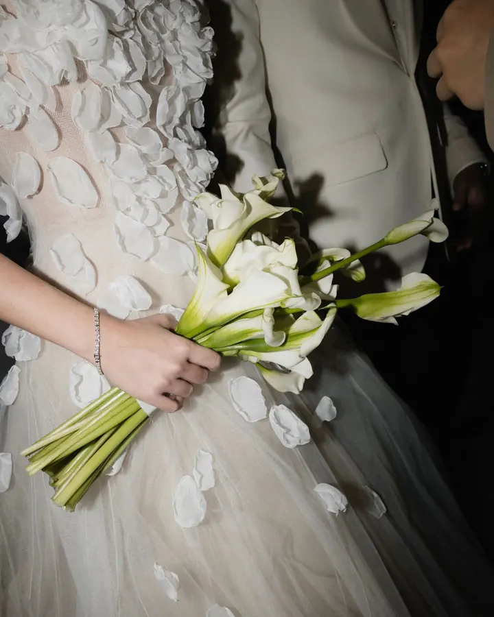 Close-up of a tulle gown covered in fabric petals, the bride holding white calla lilies beside her groom