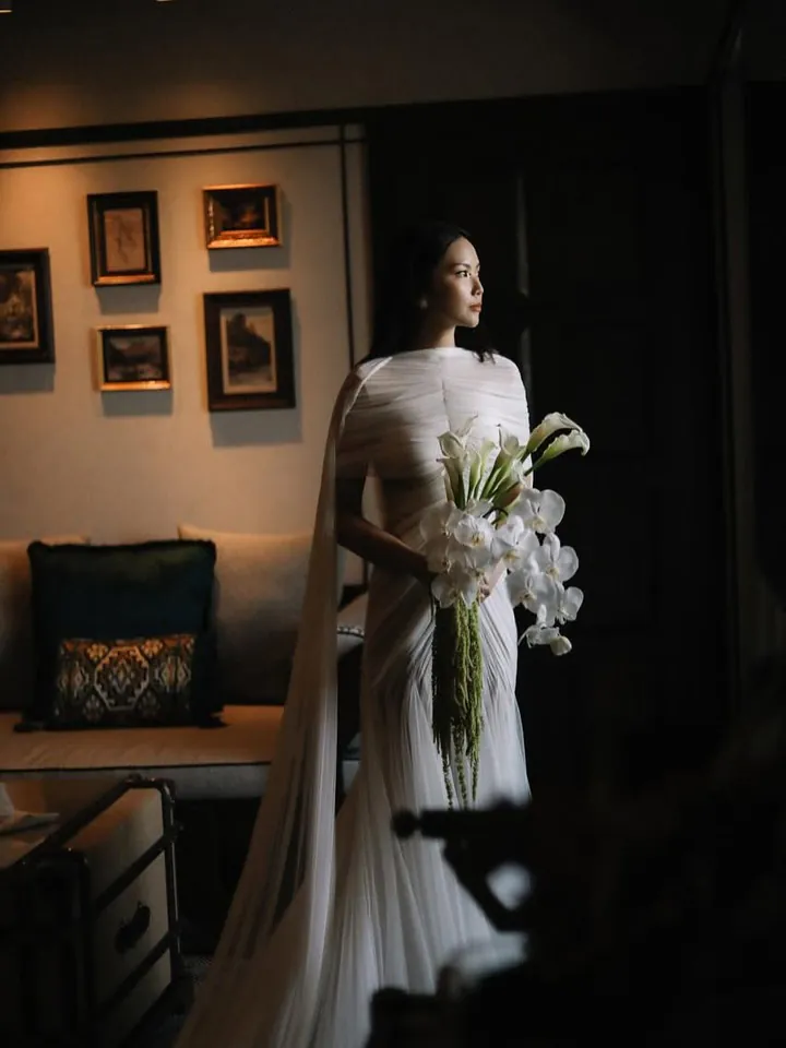 A bride holding a long cascade of white calla lilies in a warmly lit room hung with framed pictures