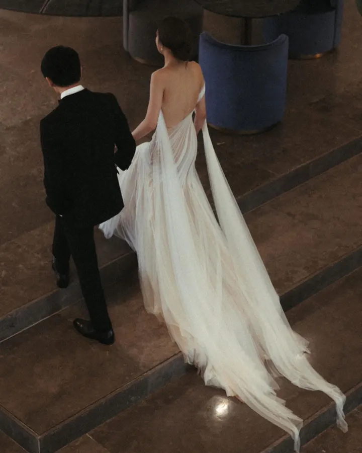 A bride and groom walking down steps, her backless gown trailing a long sheer cape