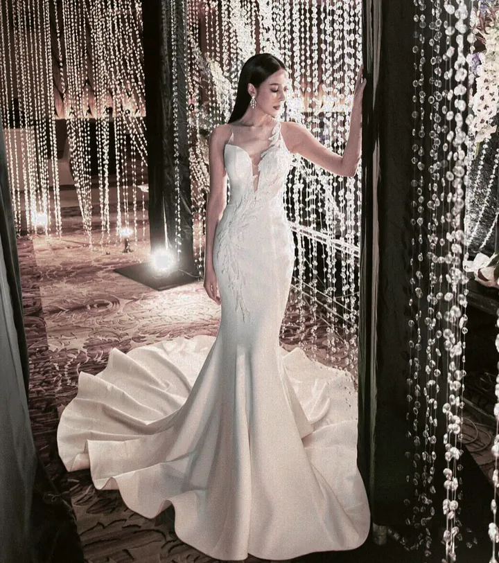 A bride in a satin mermaid gown with an embellished bodice, framed by strands of hanging crystals, her train pooled on the floor