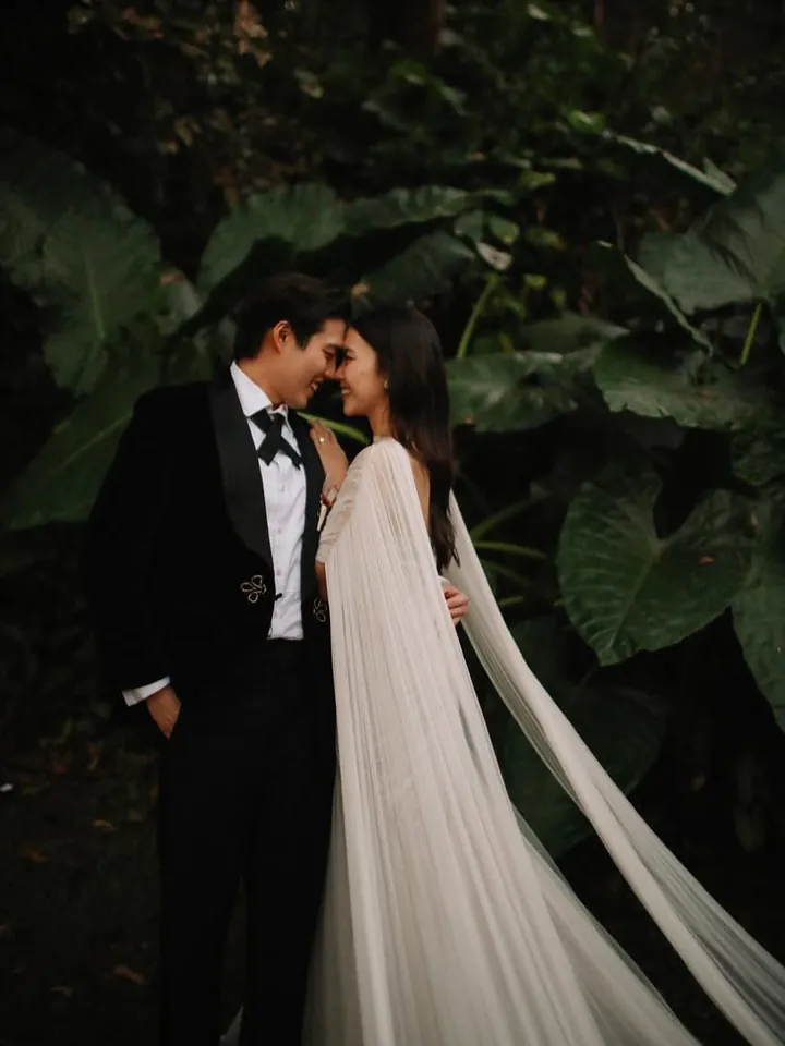 A bride and groom touching foreheads among tropical leaves, her long sheer cape falling to the ground