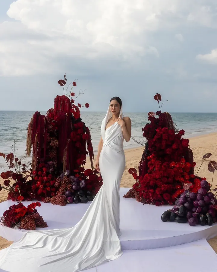 A bride in an ivory satin mermaid gown with a long train on a white platform by the sea, between tall arrangements of deep red flowers
