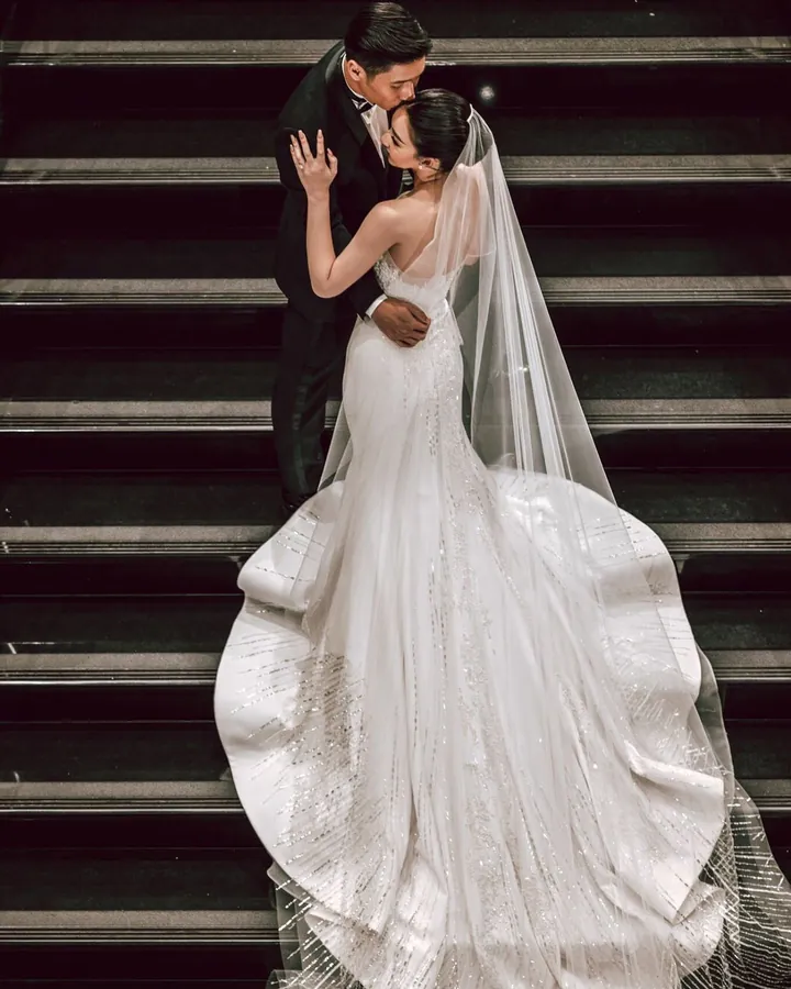 A groom kissing a bride on red-carpeted stairs, her lace mermaid gown and veil spreading into a wide scalloped train