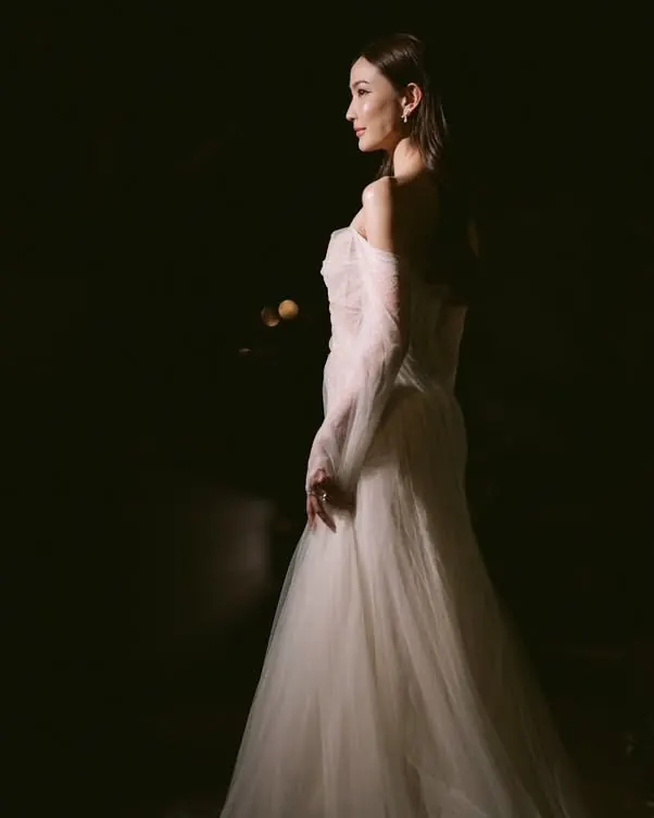 A bride in profile in a dark room, an off-shoulder sheer tulle gown catching a single light