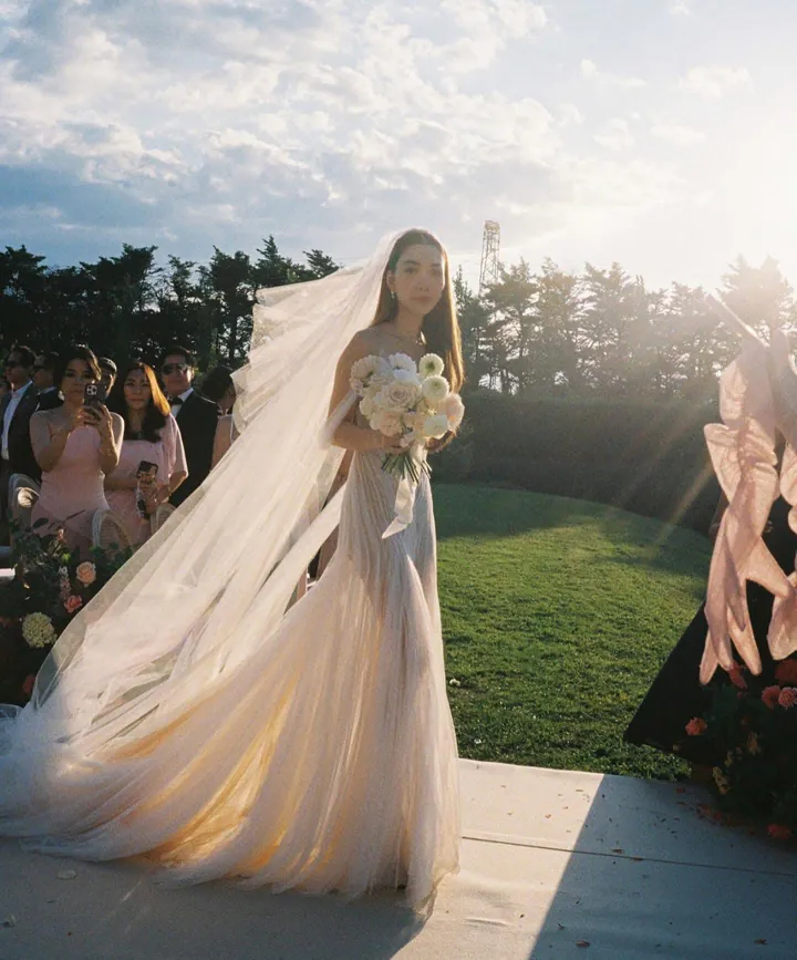 A bride with a long sheer veil holding white flowers in a garden at golden hour, guests behind her