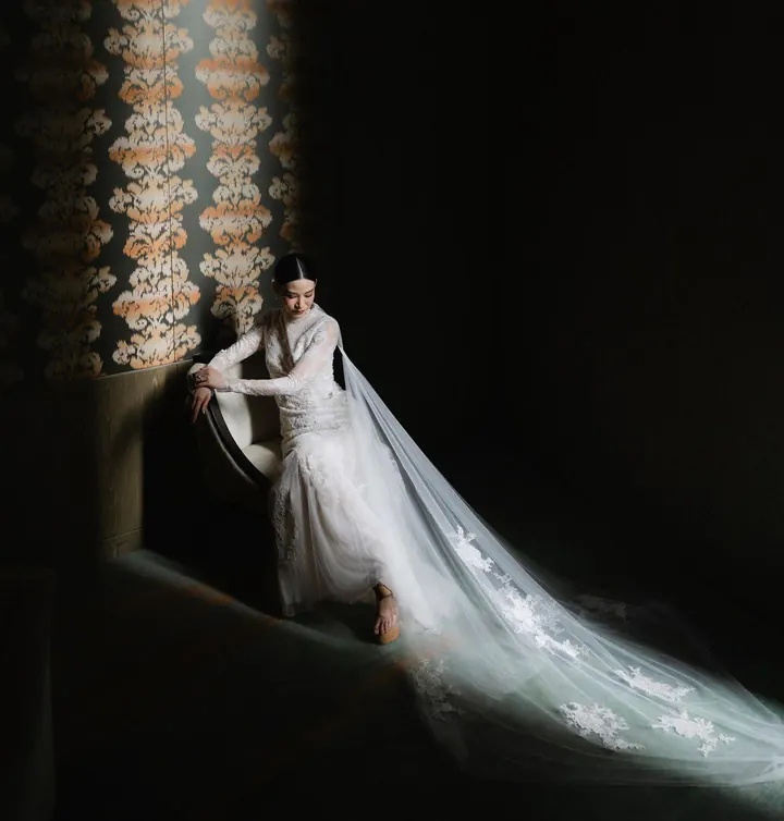A bride seated in a dark room, her long lace-appliqué train spread across the floor in a shaft of light