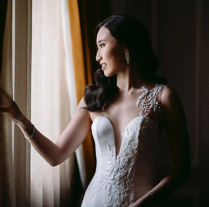 A bride at a curtained window in a gown with a plunging neckline and lace appliqué across the bodice