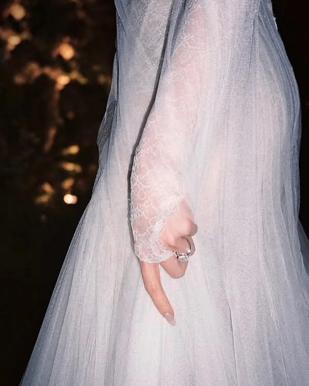 Close-up of a sheer lace sleeve ending at the hand, a ring on one finger, warm lights behind