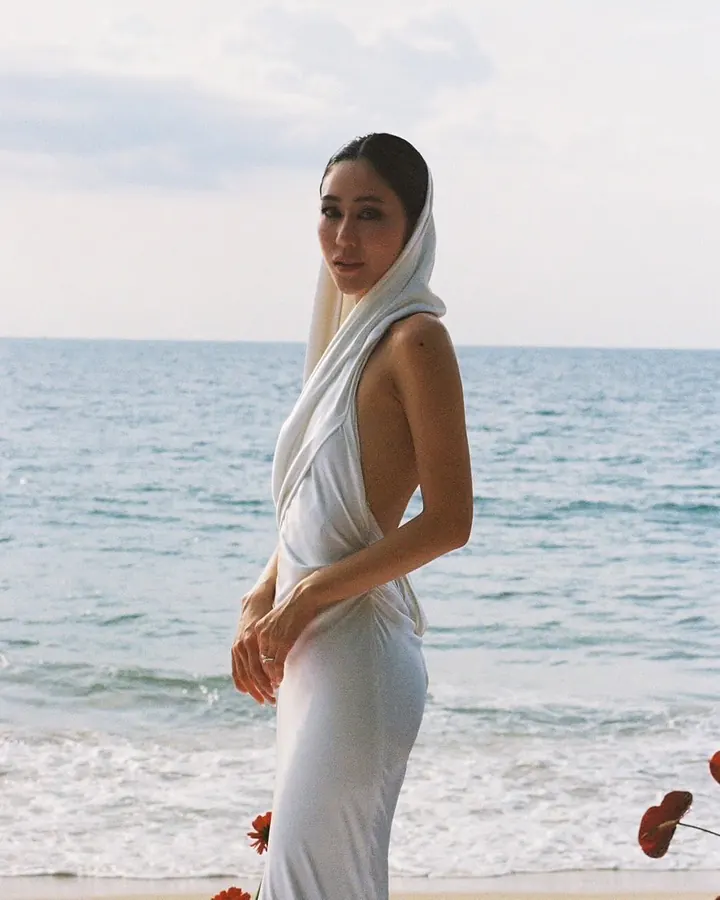 A woman on a beach in a white hooded, backless gown of fluid draped fabric