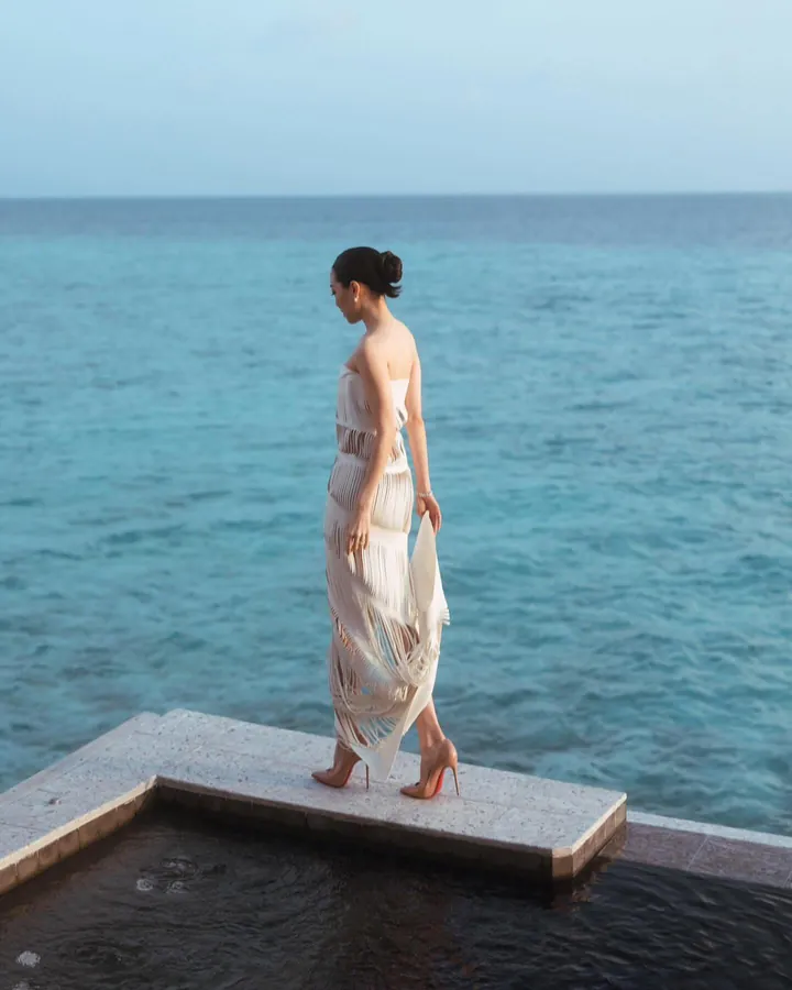 A woman in an ivory cut-work dress walking along the edge of a pool above a turquoise sea